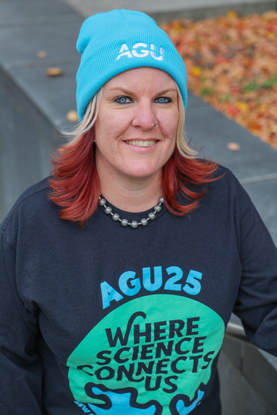 A woman wears the long-sleeve AGU25 t-shirt, with the Earth and conference theme printed in blue and green on black, and a bright blue beanie with the AGU logo