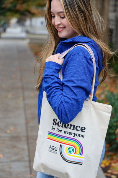 A woman wears a blue lightweight hoodie with the AGU logo printed in white and holds a cotton tote bag printed with the slogan "Science is for everyone"