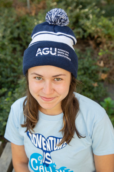 A woman wears the AGU pom-pom beanie. She also wears a light blue t-shirt featuring a whale's tail and ocean wave, plus "Adventure is Calling," all printed in blue and white.