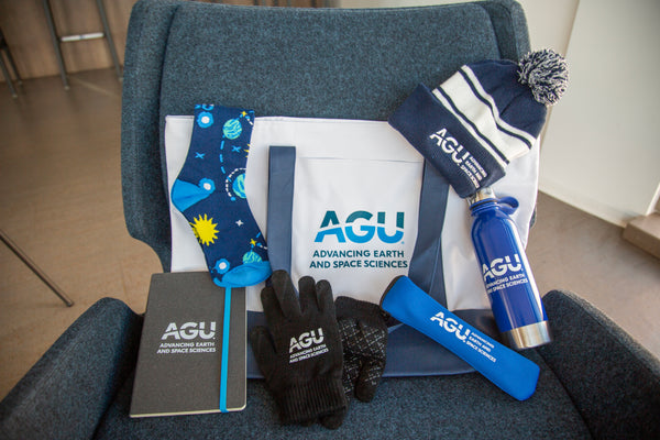 A dark grey notebook with a blue strap and the AGU logo is displayed next to other merchandise, including a water bottle, gloves, tote bag, utensil set and beanie.