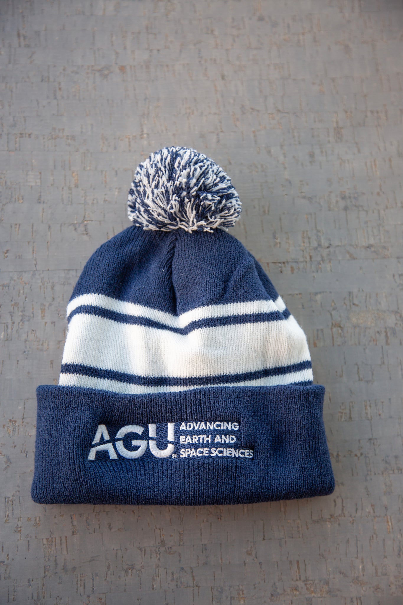 A navy blue and white beanie is displayed. It has stripes, a pom-pom and the AGU logo embroidered in white on a navy background.